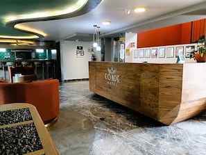 Un comptoir d’accueil d’hôtel avec un revêtement en bois et l’inscription « CONDE HOTEL ». Des tableaux encadrés sont accrochés au mur derrière le comptoir.