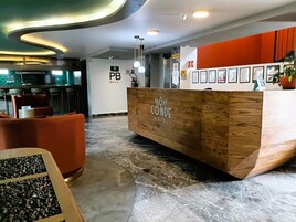 Un mostrador de recepción de hotel con detalles de madera y el nombre 'CONDE HOTEL' visible. En la pared detrás del mostrador hay fotografías enmarcadas.