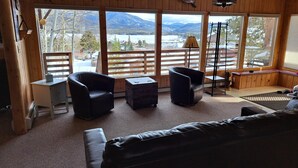 L’intérieur de ce chalet accueillant est doté d’une grande fenêtre offrant une vue panoramique sur les montagnes et un lac. On y trouve deux fauteuils en cuir noir, une petite table et un canapé.