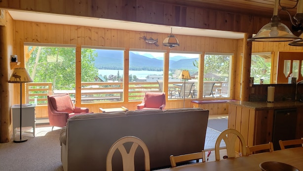 Une cabane en bois comprenant un coin repas, une cuisine et de grandes fenêtres donnant sur un lac et des montagnes.