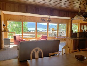 A wooden cabin with a dining area, a kitchen, and large windows offering a view of a lake and mountains.