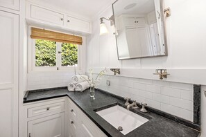 Een badkamer met een wit wastafelmeubel, een wastafel, een spiegel en een raam met uitzicht op groen.