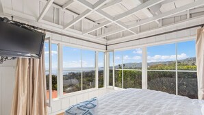 A bedroom with a large bed, a television, and multiple windows offering a view of the landscape. - Charming Cottage with Beautiful Ocean Views (Laguna Beach)