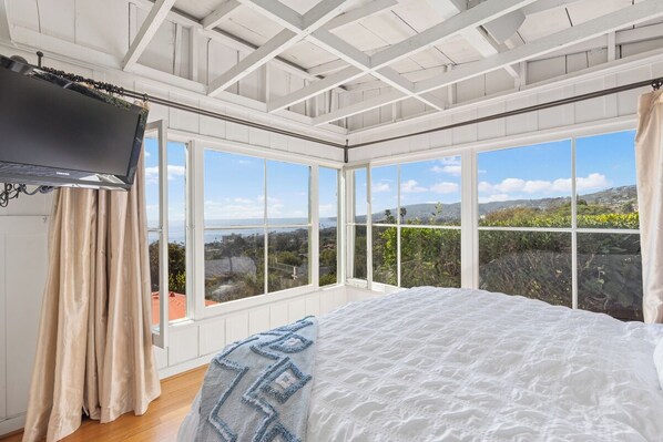 A bedroom with a large bed, a television, and multiple windows offering a view of the landscape. - Charming Cottage with Beautiful Ocean Views (Laguna Beach)