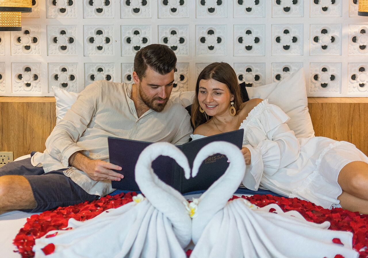A couple sitting on a bed with a heart-shaped arrangement of towels and rose petals.