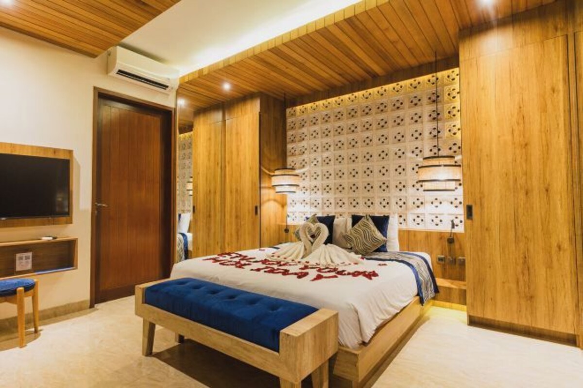 A bedroom with a wooden ceiling, a bed with a blue upholstered bench, a wall-mounted TV, and a patterned headboard.