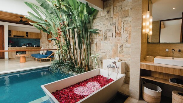 A modern bathroom with a stone wall, a bathtub filled with red pebbles, a wooden vanity, and a large plant. -  The Mavila Sanur (Denpasar)