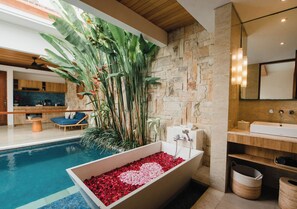 A modern bathroom with a stone wall, a bathtub filled with red pebbles, a wooden vanity, and a large plant. -  The Mavila Sanur (Denpasar)