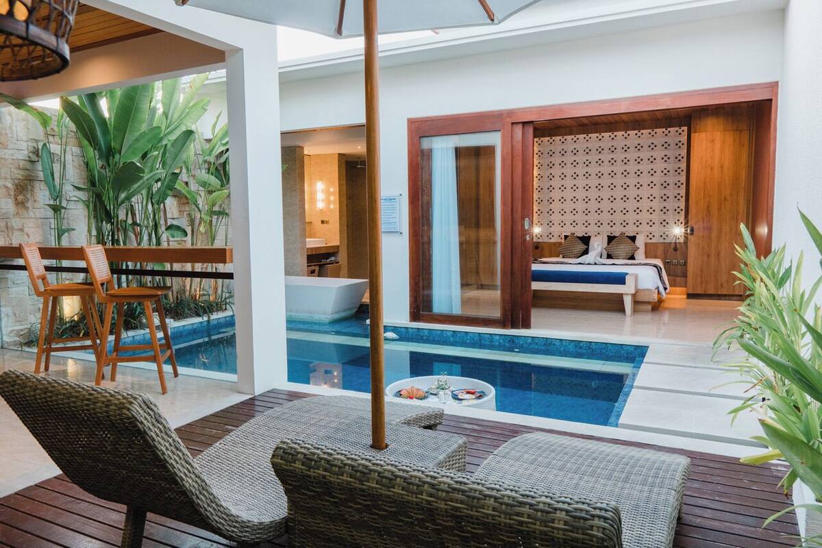 Zona junto a la piscina con mobiliario de mimbre, una piscina pequeña y vista a un dormitorio con cama y puerta de madera.