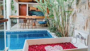 A modern bathroom with a large bathtub filled with rose petals, a stone wall, and a view of a dining area with a pool. -  The Mavila Sanur (Denpasar)