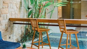 A modern room with a stone wall, a blue sofa, two wooden chairs, a small round table with a decorative item, and a view of a pool through a glass door. -  The Mavila Sanur (Denpasar)