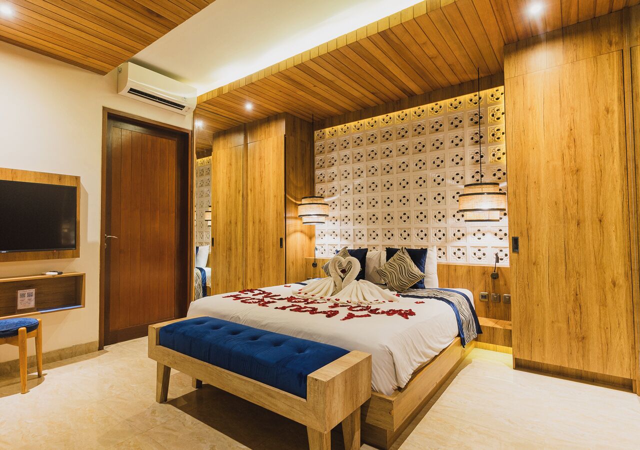 Un dormitorio con techo de madera, una cama grande con un banco tapizado azul, un televisor montado en la pared y un panel decorativo.