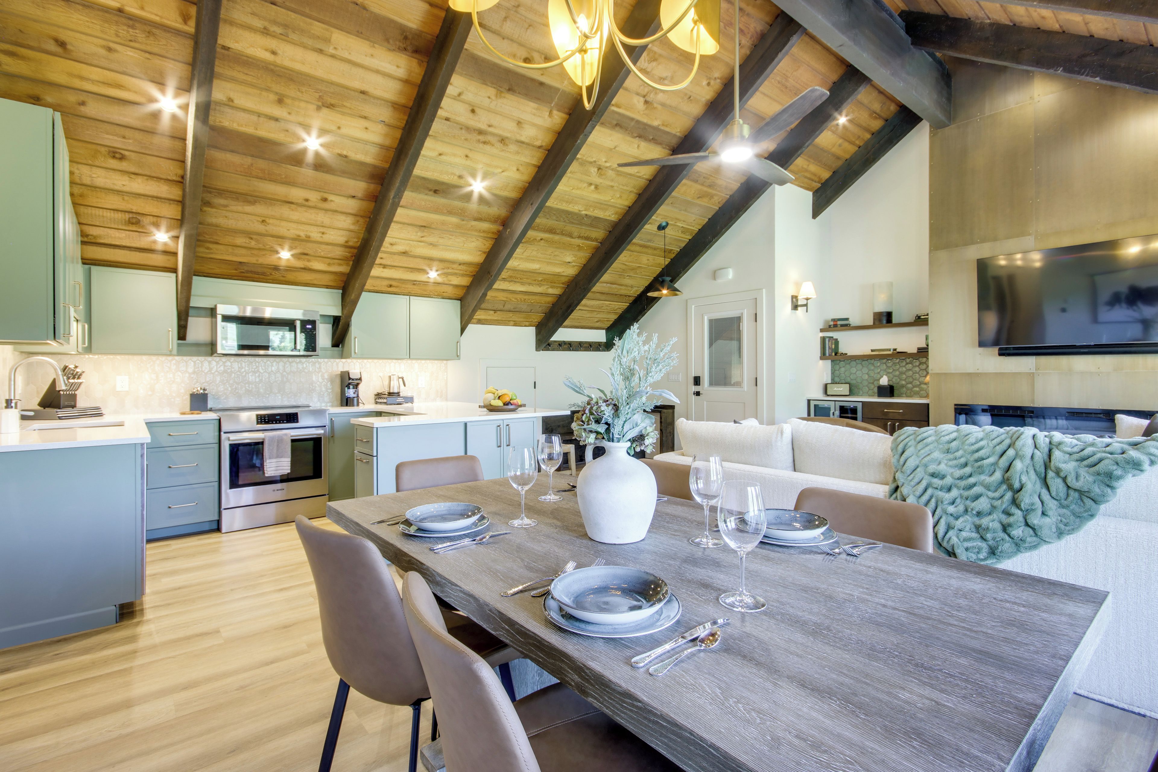 Une cuisine moderne avec un plafond en bois, un coin repas avec une table en bois mise pour deux personnes, et un salon avec un canapé et un téléviseur.