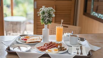 Bandeja de café da manhã com torradas, ovos, bacon, cogumelos e um copo de suco de laranja sobre uma mesa com um vaso de flores branco.
