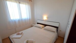 A neatly made bed with pillows and a bedside table, a wall-mounted light, and a window with sheer curtains. - Apartment Marinela 1 (Njivice)