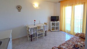 A living room with a sofa, a TV on a stand, a dining table with chairs, a clock on the wall, and a window with curtains. - Apartment Marinela 1 (Njivice)