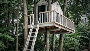 Een houten boomhut met een ladder, omgeven door weelderige begroeiing.