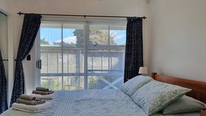 A bedroom with a bed, a nightstand, a window with curtains, and a view of the outside. - Poripori Homestead (Lower Kaimai)