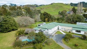 A large, single-story building with a green roof, surrounded by lush greenery and hills. - Poripori Homestead (Lower Kaimai)