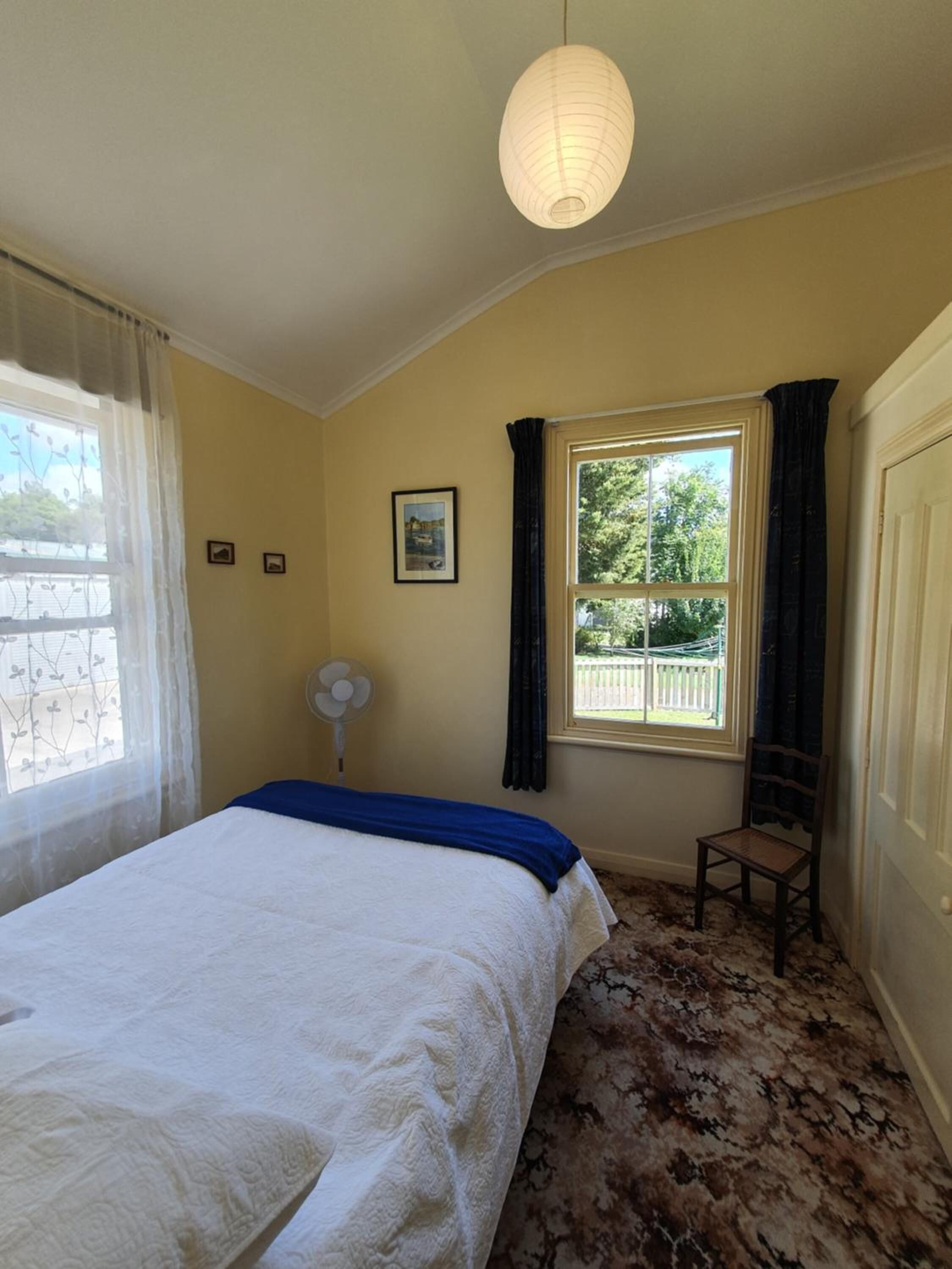 A bedroom with a bed, a window with curtains, a chair, a fan, and a picture on the wall.