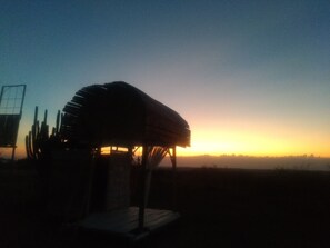 Una estructura de madera rústica con techo de paja, posiblemente un pabellón o un pequeño refugio, rodeada de cactus, con un cielo al atardecer.