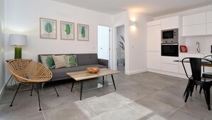 Un salon moderne avec un canapé gris, un fauteuil en osier, une table en bois sur laquelle se trouve un saladier de fruits, et un coin cuisine doté de meubles blancs et d’appareils électroménagers.