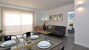 Un salon moderne avec une table à manger en verre pour deux personnes, un canapé et une fenêtre équipée de stores.