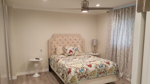 Une chambre à coucher avec une tête de lit capitonnée, une couvre-lit à motifs floraux, une lampe de chevet et un ventilateur de plafond.