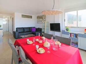 Un salón moderno con una mesa de comedor roja dispuesta para una comida, un sofá negro, un televisor de pantalla plana y un ventanal con vistas.