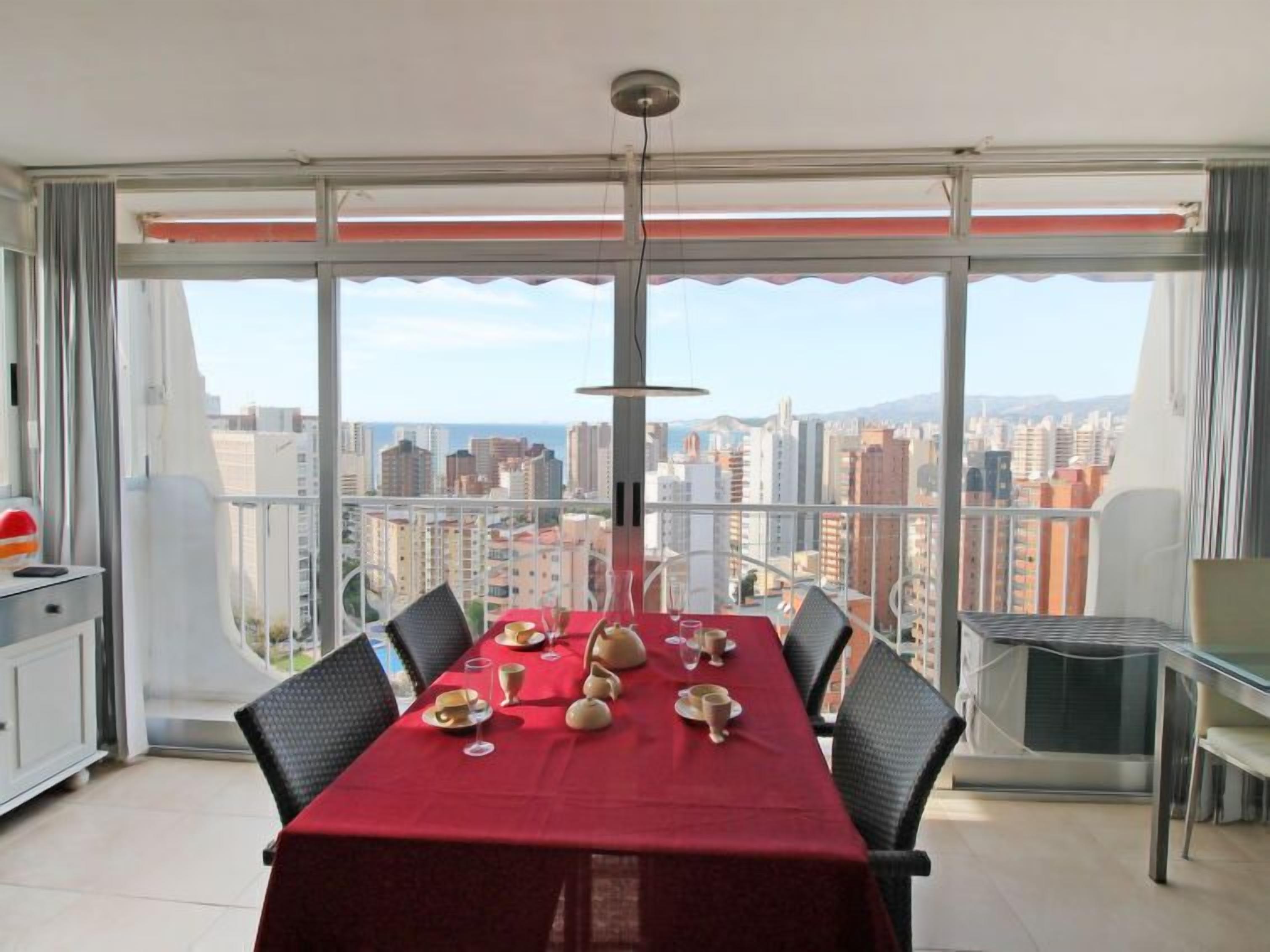 Ein Essbereich mit einem roten Tisch, gedeckt für sechs Personen, großen Fenstern mit Stadtblick und einem Balkon mit einem Stuhl.