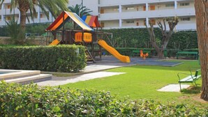 A playground with slides and climbing structures, surrounded by well-maintained grass and hedges, with a multi-story building in the background. - Park Playa Levante by Interhome (Benidorm)