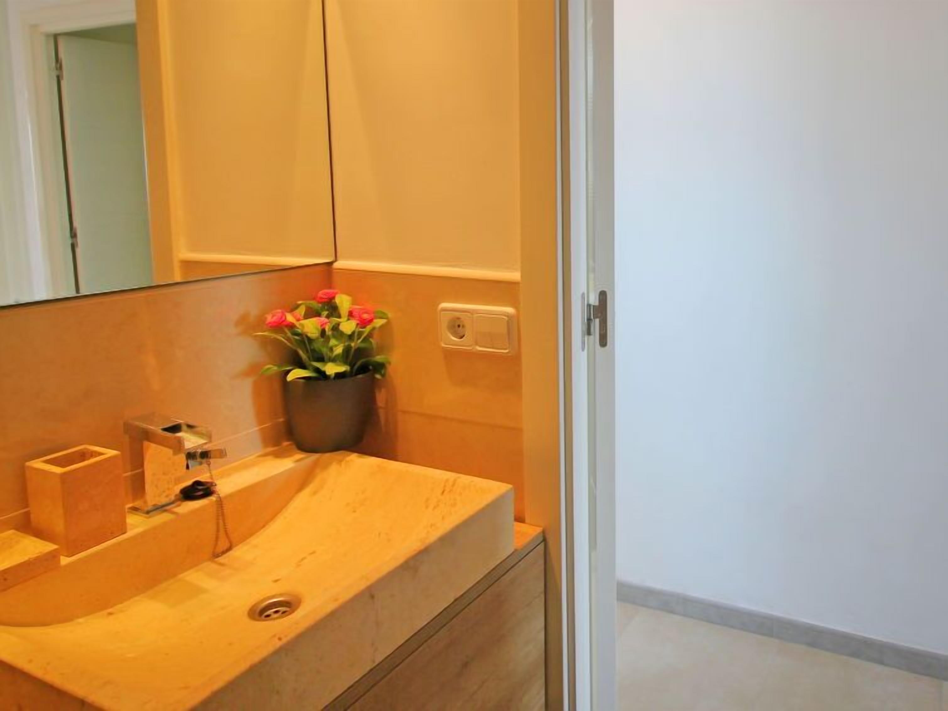 Une salle de bain avec un meuble-lavabo en bois, un évier, un miroir et une plante en pot aux fleurs rouges.