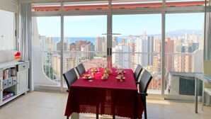 A dining area with a red table set for four, large windows offering a city view, and a small bookshelf to the left. - Park Playa Levante by Interhome (Benidorm)