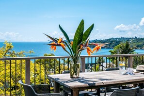 A wooden table with chairs on a balcony overlooking a scenic view of the sea and a distant town. - 16 Waikare | You've Got It Maid (Oneroa)
