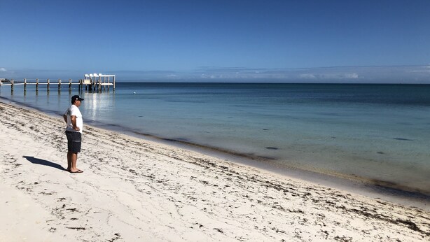Ein Strand mit einem Steg, der sich in ruhiges Wasser erstreckt, bewölkter Himmel und eine Person, die am Sand steht.