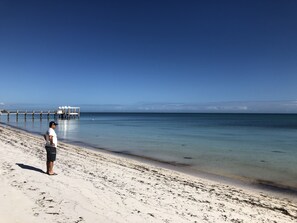 A beach with a pier extending into calm waters, a clear sky, and a person standing on the sand.