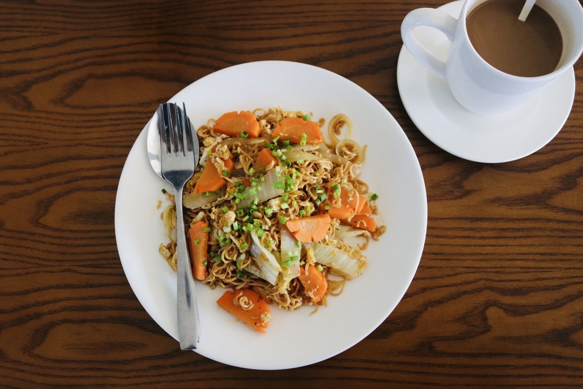 A plate of noodles with vegetables and a cup of coffee on a wooden table.