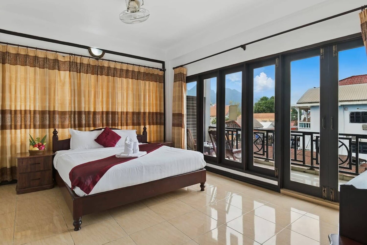 Habitación de hotel con una cama grande, una mesita de noche, un ventanal con vistas a edificios y montañas, y un balcón con barandilla.
