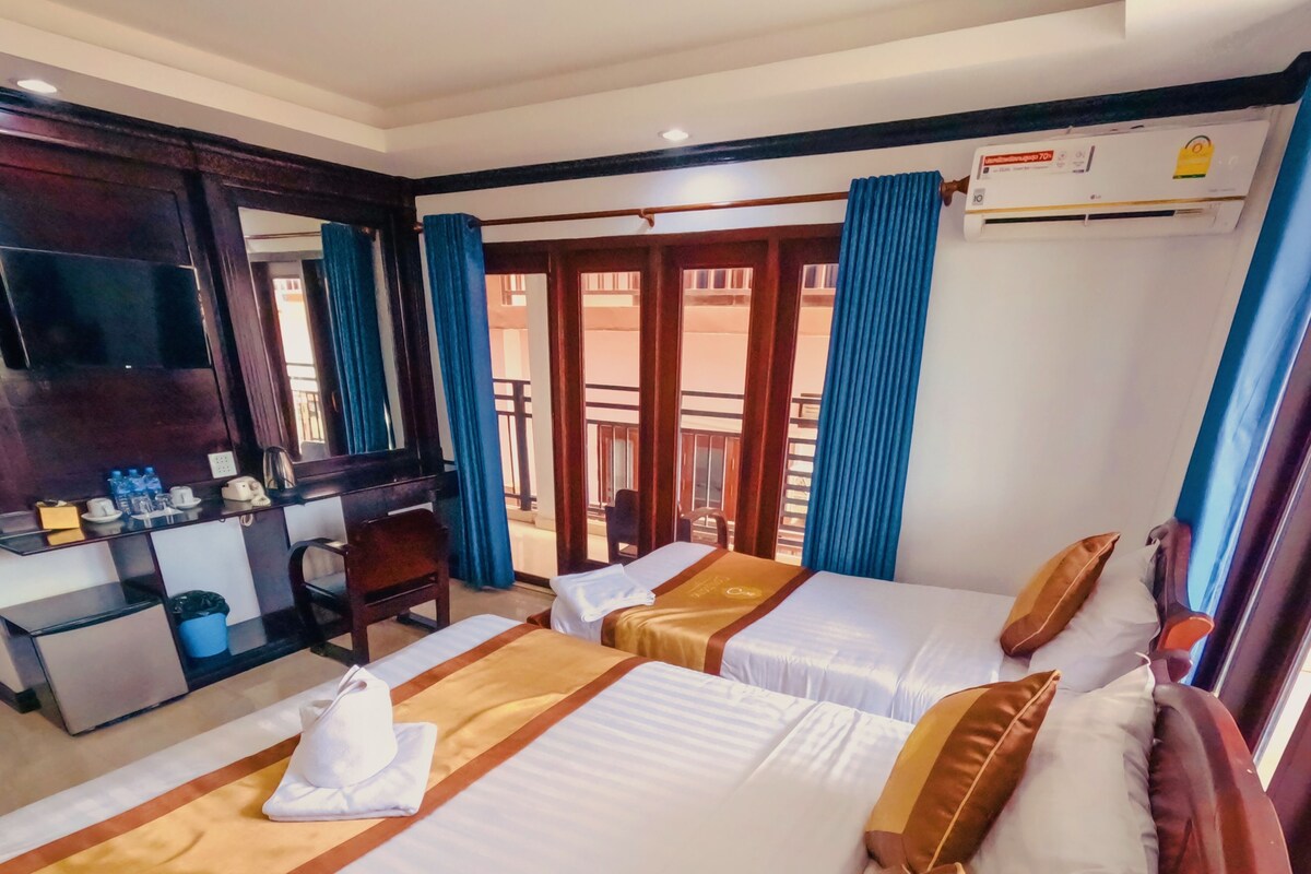 Habitación de hotel con dos camas, un escritorio con teléfono y hervidor, una silla y un balcón con cortinas azules.
