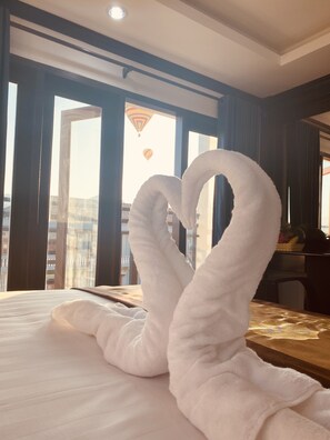 Una habitación de hotel con una cama, un arreglo de toallas con forma de corazón, una ventana con vistas a edificios y un globo aerostático en el cielo.