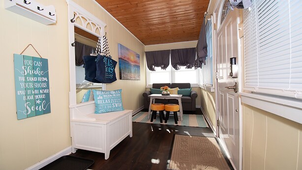 Una habitación acogedora con techo de madera, un banco blanco, una mesita con un florero y una puerta con persianas.