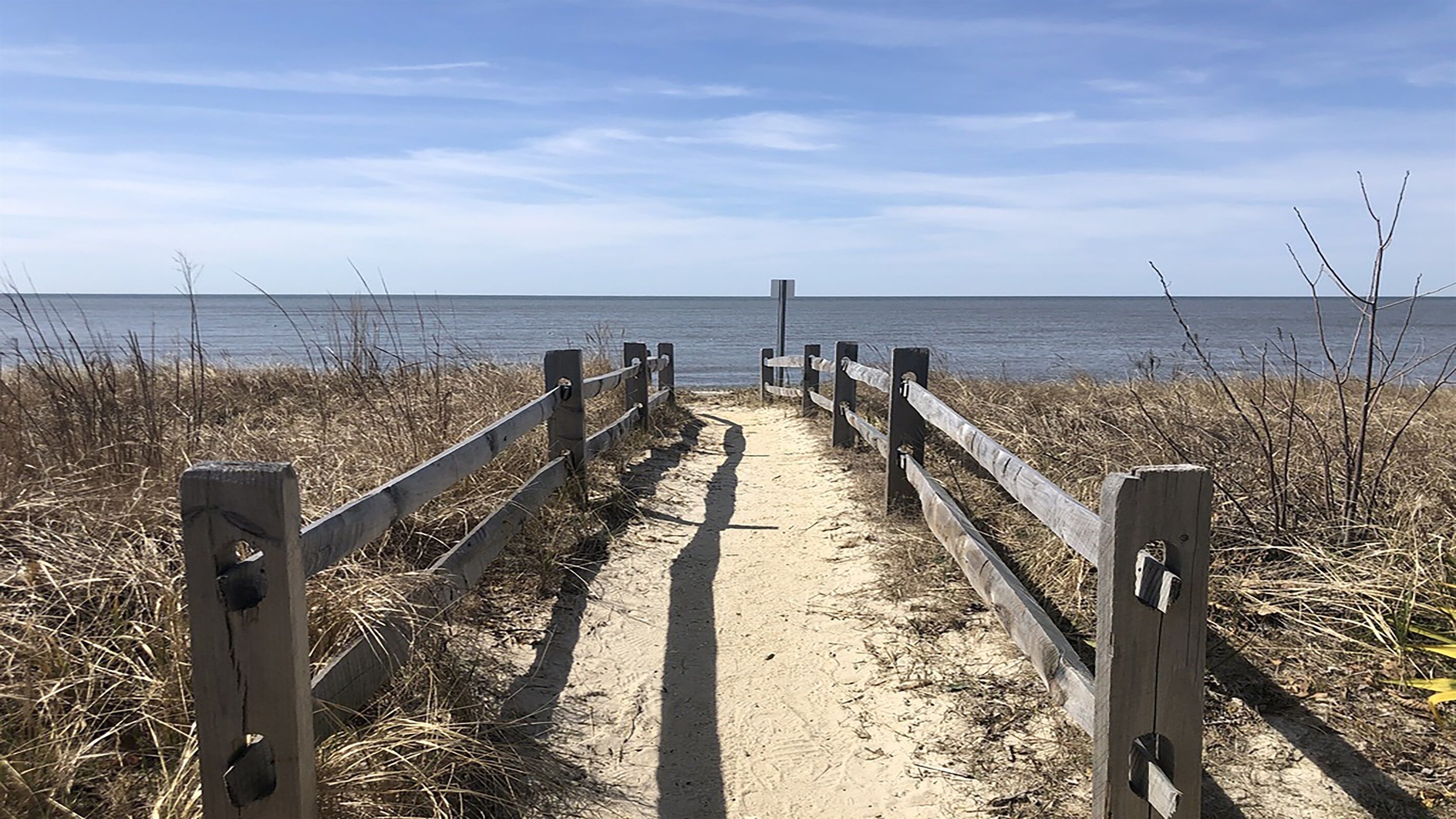 Un sentier de sable qui mène à l’eau, bordé de clôtures en bois et d’herbes sèches.