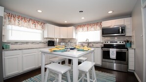Cocina moderna con armarios blancos, un microondas de acero inoxidable y un horno. Hay una mesa de comedor con sillas y una alfombra a rayas.