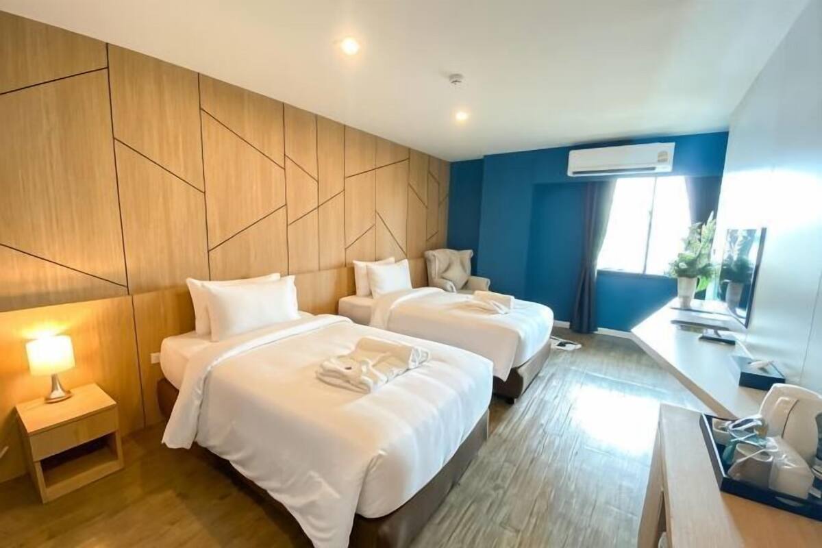 A hotel room with two beds, a desk, a chair, a lamp, and an air conditioning unit.