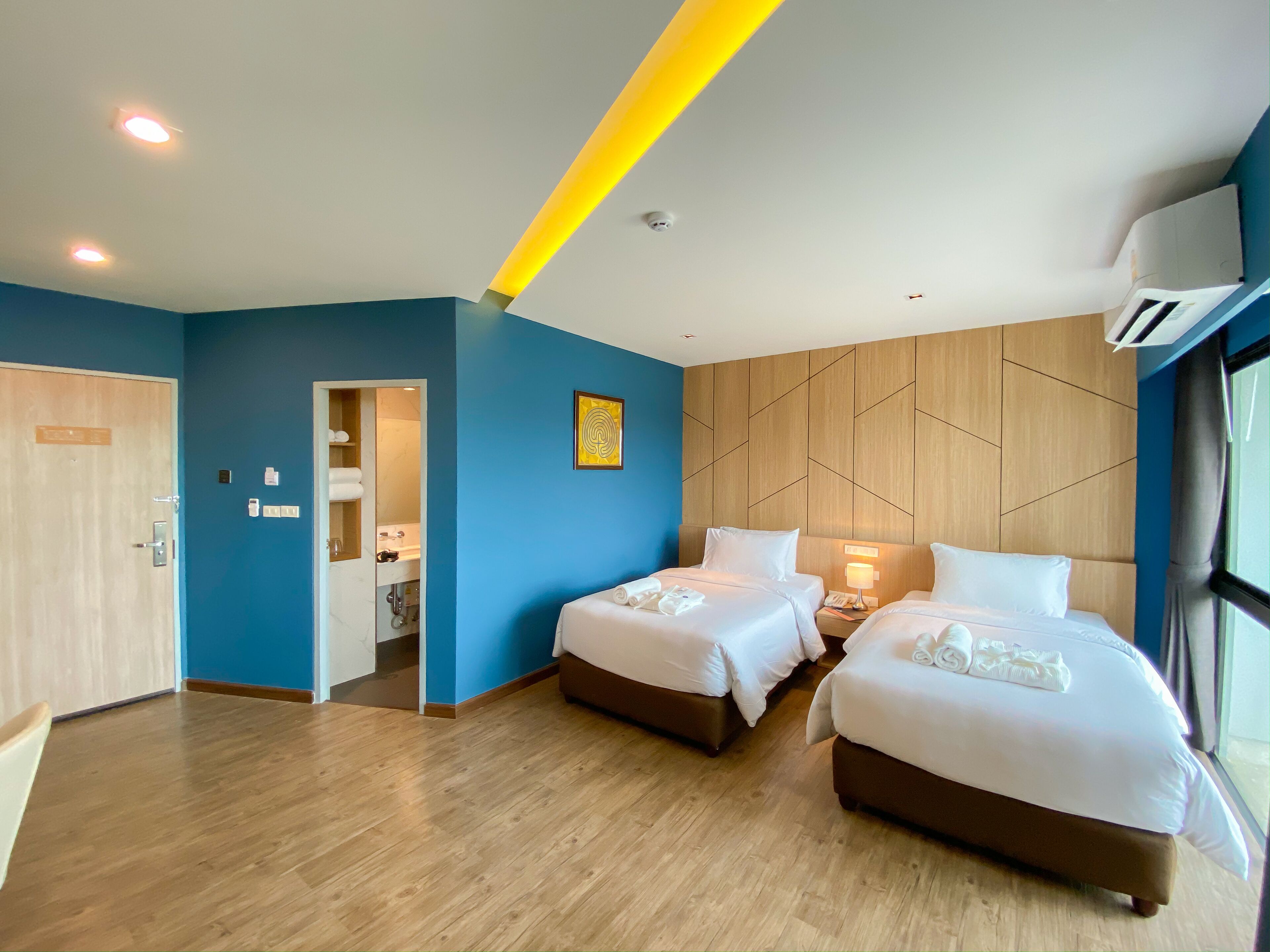 A hotel room with two beds, a wooden floor, a blue accent wall, and a large window.