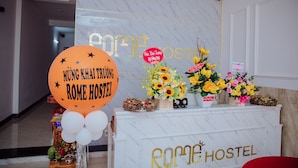 A reception area with a marble wall, a white reception desk, and a sign that reads "Rome Hostel." There are flowers and a balloon with text on it. - Rome Hostel (Tuy Hoa)