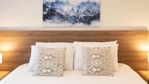 A hotel room with a wooden headboard, a bed with white linens, two patterned pillows, a wall-mounted air conditioner, and a painting of a mountain landscape. - Blacktown Tavern (Blacktown)