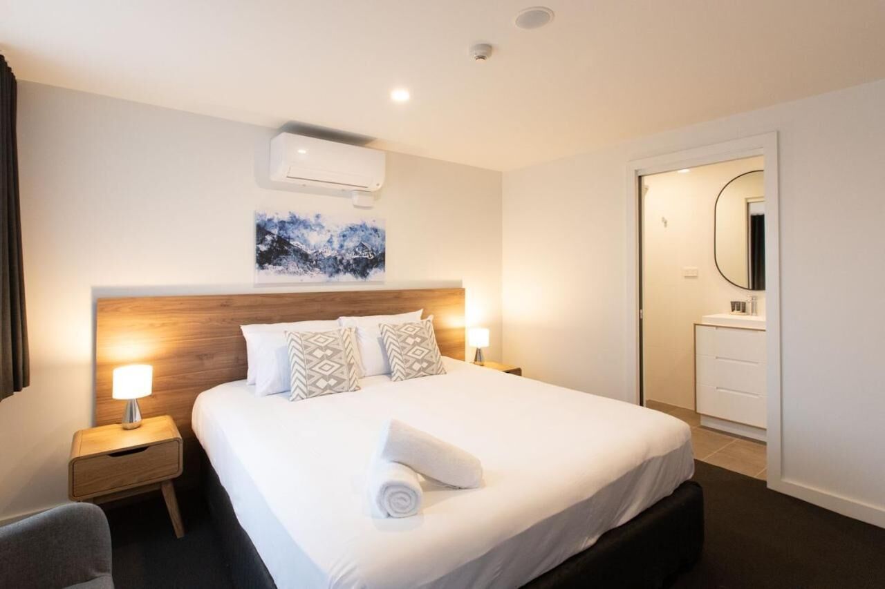Habitación de hotel con una cama grande, dos lámparas de noche, un cabecero de madera, aire acondicionado instalado en la pared, un cuadro, y un baño con espejo y una toalla sobre la cama.