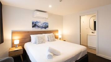 Habitación de hotel con una cama grande, dos lámparas de noche, un cabecero de madera, aire acondicionado instalado en la pared, un cuadro, y un baño con espejo y una toalla sobre la cama.