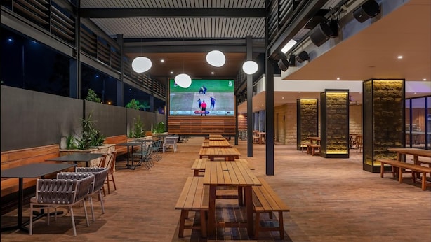 A spacious indoor dining area with wooden tables and chairs, a large screen displaying a sports event, and a modern architectural design with exposed beams and pendant lighting. - Blacktown Tavern (Blacktown)